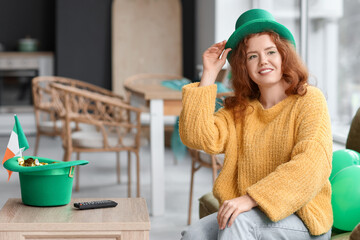 Young redhead woman in Leprechaun's hat sitting on sofa at home. St. Patrick's Day celebration