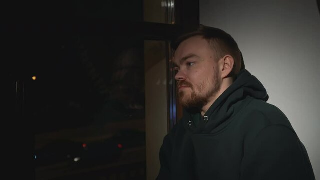 Young man looks depressingly out night window. Media. Lonely man looks dejectedly out window of house. Depressed man looks out window