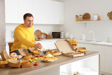 Young man at table full of unhealthy food in kitchen. Overeating concept