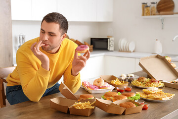 Young man with doughnut licking fingers at table full of unhealthy food in kitchen. Overeating...