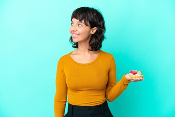 Young mixed race woman holding a tartlet isolated on blue background looking side