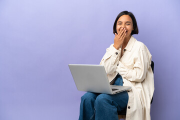 Young mixed race woman sitting on a chair with laptop isolated on purple background happy and smiling covering mouth with hand