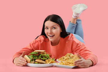 Young woman with unhealthy food lying on pink background. Overeating concept