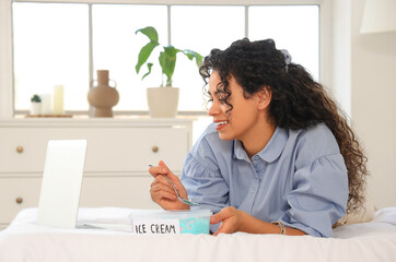 Beautiful young African-American woman with basket of ice cream and modern laptop lying on bed at...