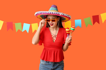 Happy young woman in Mexican sombrero hat and with maracas on orange background