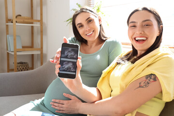 Young pregnant lesbian couple holding mobile phone with sonogram image at home