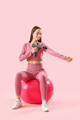 Sporty young woman massaging her arm with percussive massager on fitball against pink background
