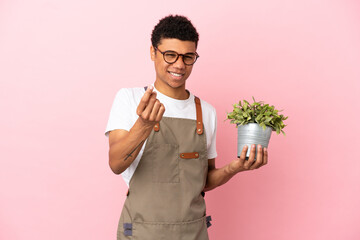 Gardener African man holding a plant isolated on pink background making money gesture