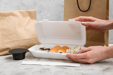 Female hands holding plastic container with tasty sushi rolls, chopsticks and paper bags on light...