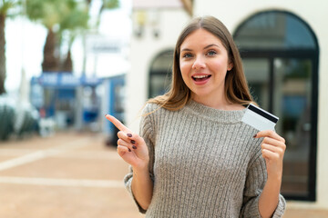 Young pretty blonde woman holding a credit card at outdoors surprised and pointing finger to the...