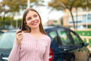 Young pretty blonde woman holding car keys at outdoors looking up while smiling