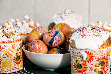 Easter cakes with protein glaze, sugar sprinkles, marshmallow on top and eggs decorated with patterns in a plate, on a with blurred background. Orthodox Easter in eastern Europe.