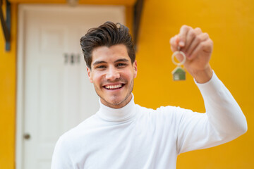 Young caucasian man at outdoors holding home keys with happy expression