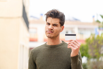 Young caucasian man holding a credit card at outdoors with sad expression