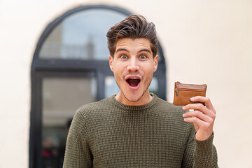 Young caucasian man holding a wallet at outdoors with surprise and shocked facial expression