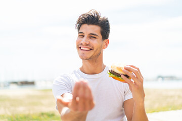 Young caucasian man holding a burger at outdoors inviting to come with hand. Happy that you came