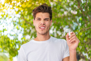 Young caucasian man holding invisible braces at outdoors with sad expression