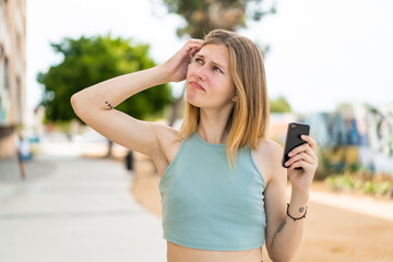 Young blonde woman using mobile phone at outdoors having doubts and with confuse face expression