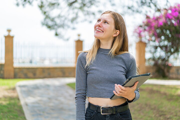 Young blonde woman holding a tablet at outdoors looking up while smiling