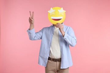 Man holding emoticon with stars instead of eyes and showing peace sign on pink background