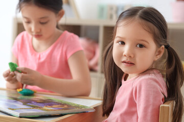 Obraz premium Cute little Asian happy sisters with toys reading book at table in child room
