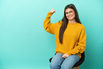 Young caucasian woman sitting on a chair isolated on blue background doing strong gesture