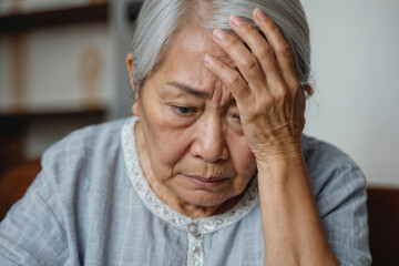 Fototapeta premium A gray-haired Asian woman pensioner hold his head in sadness, she sits at a table against the backdrop of a blurry, light hall