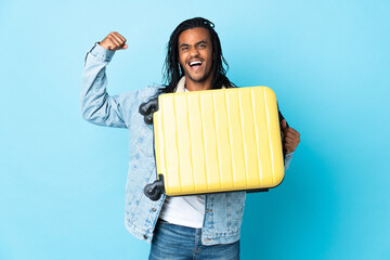 Young African American man with braids isolated on blue background in vacation with travel suitcase