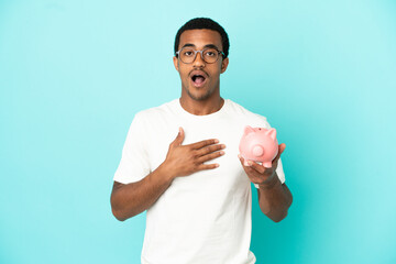 African American handsome man holding a piggybank over isolated blue background surprised and...