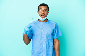 African American dentist holding tools over isolated blue background with surprise facial expression