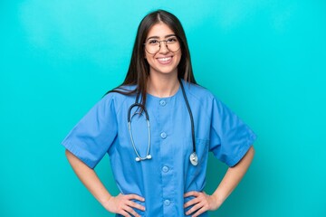 Young nurse caucasian woman isolated on blue background posing with arms at hip and smiling