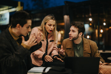 Three young business professionals in a lively discussion over a laptop at a cafe during nighttime.