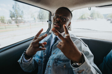 A trendy young black businessman flashing peace signs while sitting in the back of a car, dressed...