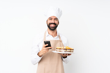 Young man holding muffin cake over isolated white background sending a message with the mobile