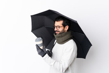 Caucasian handsome man with beard holding an umbrella and a coffee to take away over isolated white wall