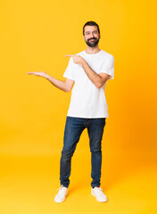 Full-length shot of man with beard over isolated yellow background holding copyspace imaginary on...