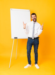 Full-length shot of businessman giving a presentation on white board over isolated yellow background making rock gesture