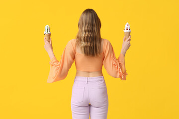 Young woman holding ice creams in waffle cone on yellow background, back view