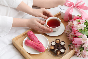 Tasty breakfast served in bed. Woman with tea, desserts, gift box, flowers and I Love You card at home, closeup