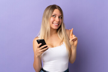Young blonde woman using mobile phone isolated on purple background with fingers crossing