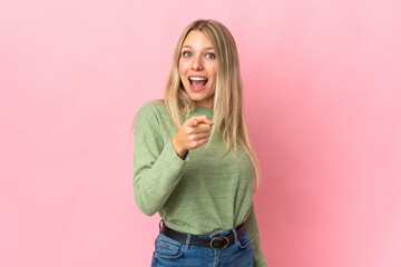 Young blonde woman isolated on pink background surprised and pointing front
