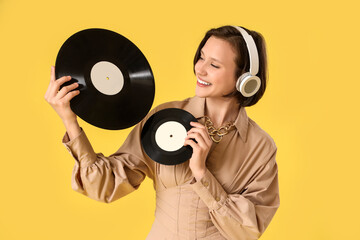 Beautiful young woman in headphones with vinyl disks on yellow background
