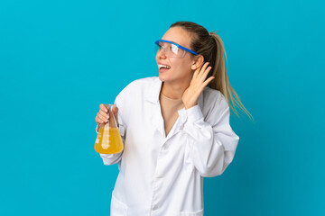 Young scientific woman isolated on blue background listening to something by putting hand on the ear