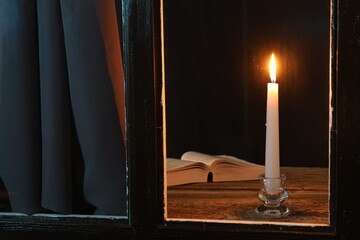 Burning candle and Bible on wooden table at night, view through window