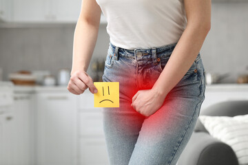 Woman holding sticky note with drawn sad face and suffering from cystitis symptoms indoors, closeup