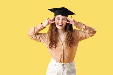 Happy female student in graduation hat showing victory gesture on yellow background