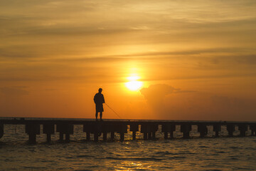 silhouette of a person fishing at the pier on the beach with sunset behind