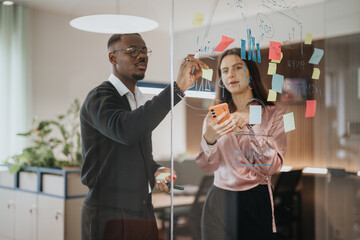 Two multiracial young business professionals collaborate in a brightly lit office, using sticky...