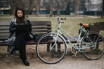 Young and attractive businesswoman in stylish outfit focused on work outdoors near her classic bicycle on a bench