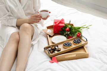 Tasty breakfast served in bed. Woman with tea, desserts, gift box and flowers at home, closeup